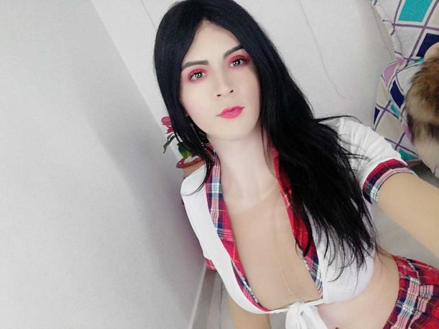 DanielaMonty with long black hair, red eyeshadow, pink lipstick, white plaid top revealing cleavage, and matching skirt, standing in a white-walled room, looking at the camera.