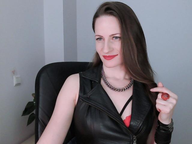 AbyX with long brown hair, red lipstick, black leather vest, red bra, chain necklace, holding red ring, sitting in black chair, smiling. Boobs visible, fair skin.