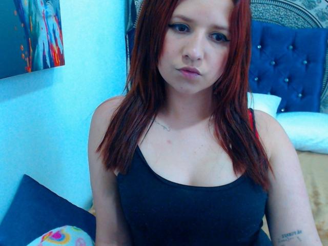 Kkatalina, com cabelo ruivo, vestindo uma regata preta, mostrando os seios, olhos azuis, lábios levemente curvados, tatuagem no braço direito, sentada na cama com cabeceira azul..