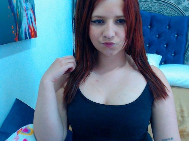 Kkatalina, con i capelli rossi, indossa una canottiera nera, mostrando piccoli seni, tatuaggi discreti e un leggero sbuffo di labbra, mentre si tiene i capelli, in una camera da letto blu..