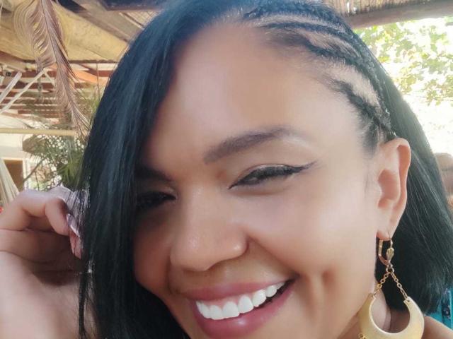 ExoticLyla's smiling face with dark braided hair, brown eyes, and black eyeliner. She's wearing gold hoop earrings, has a nose piercing, and is winking. Her skin is tan.