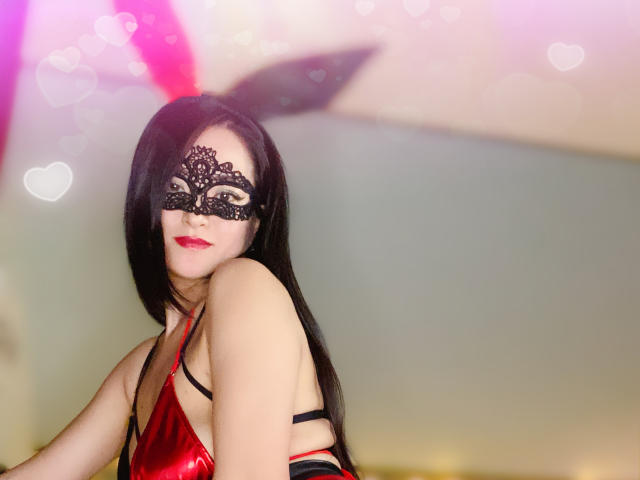 TinaAsian with long black hair, wearing a black lace mask, red shiny lingerie, and black straps, smiling with red lipstick, pink bokeh hearts in the background.