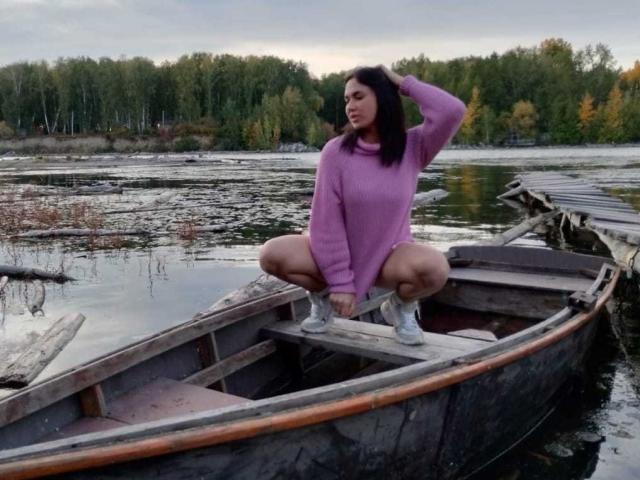 CleoLove squatting in a wooden boat, wearing a purple sweater, white sneakers, and no pants, with a hand in her hair, showing her ass and pussy.