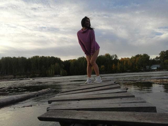 CleoLove stands on a wooden bridge, back to the camera, in a purple shirt, no pants, white sneakers, with her ass slightly raised, looking over her shoulder.