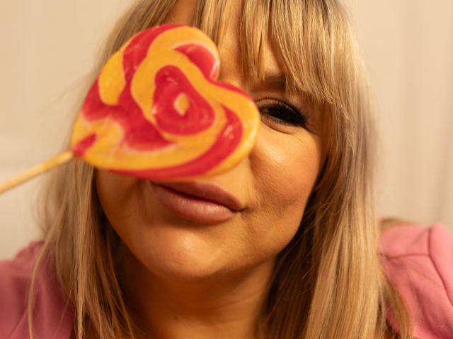 GoddessDiana with blonde hair, holding a red and yellow swirl lollipop in front of her face, slightly smiling with her lips puffed out.