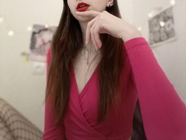 WollyMolly with long brown hair, red lipstick, and pink V-neck dress, hand near mouth, showing off small tits and delicate necklaces. Blurred white background with string lights.