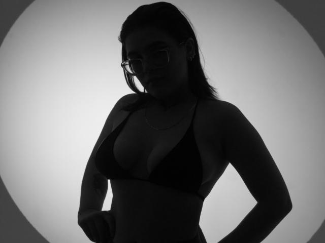 Silhouette von KimSummer mit Brille, schwarzem BH und Händen an den Hüften, die ihre Brüste und einen dezenten Hintern zeigt. Schwarz-weiß, von hinten beleuchtet, mit einer kreisförmigen Lichtquelle..