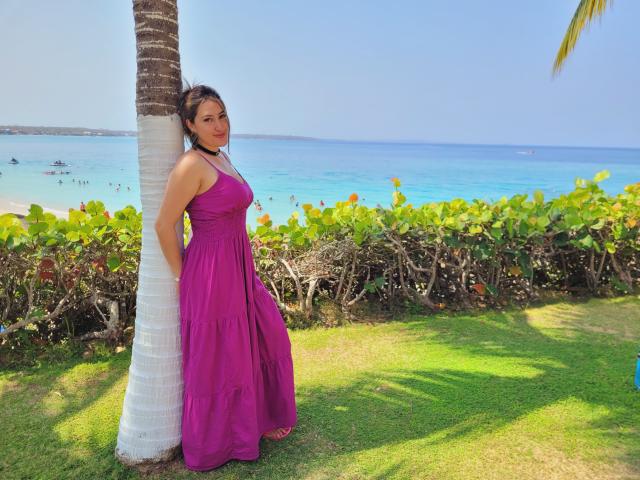 HotCarolina69 stands against a palm tree, wearing a purple dress, with her hands behind her back, looking at the camera, her ass slightly visible, by the blue ocean.