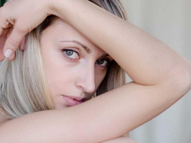 MayaUniverse with blonde hair, blue eyes, and fair skin, arms crossed over her head, looking intensely at the camera, showing a slight pout and subtle makeup.