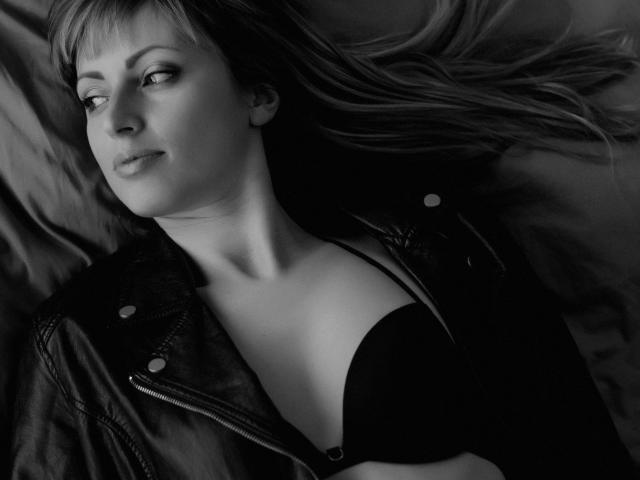 Black-and-white photo of MayaUniverse lying down with a leather jacket open, revealing her boobs in a black bra. She has long hair and a slightly annoyed expression.