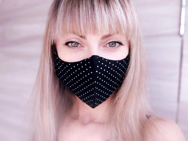 MayaUniverse with blonde bangs, wearing a black mask with white dots, covering her mouth, green eyes looking straight at the camera, light skin, and straight blonde hair.