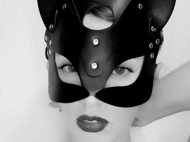 MayaUniverse wearing a black leather mask with metal studs, eyes looking sideways, dark lipstick, and her head slightly tilted, showing a sultry expression. Black and white photo.
