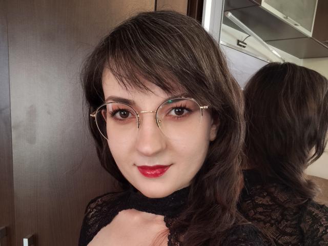 MoonBlossom with red lipstick, wearing glasses and black lace top, has wavy brown hair, fair skin, and a subtle smile. Her reflection is in the mirror.