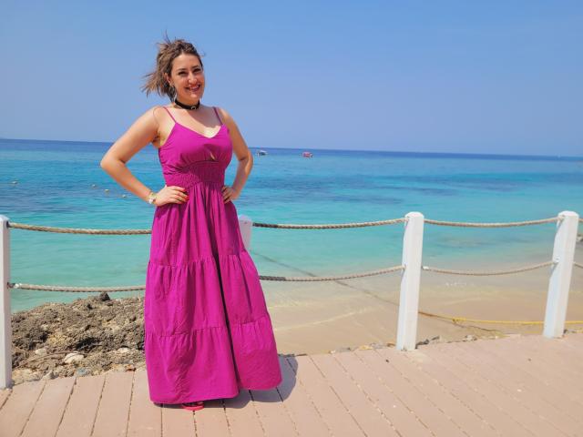 HotCarolina69 está em um vestido rosa, com as mãos nos quadris, o oceano azul ao fundo, um corrimão branco e uma corda, usando um colar preto, sorrindo, com um penteado levemente bagunçado pelo vento..