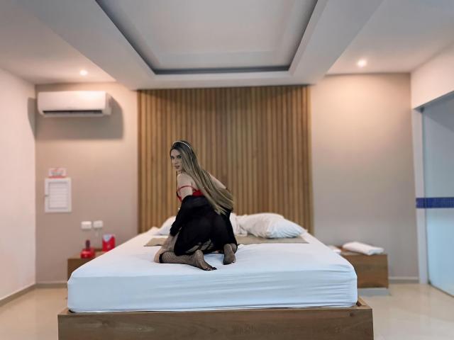 GalileaFeng with long blonde hair, red and black dress, kneeling on a white bed in a modern room, showing her bare ass, back towards camera.