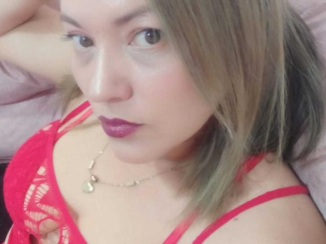 NatashaCoop, avec ses cheveux blonds, son soutien-gorge en dentelle rouge, son rouge à lèvres violet et son collier en or, regarde l'appareil photo avec un léger froncement de sourcils. Ses seins sont visibles à travers la dentelle rouge..