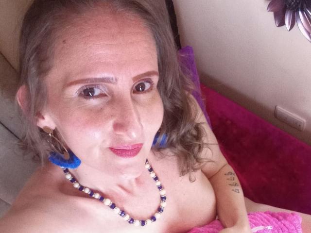 RubiAnderson with gray hair, blue feather earrings, purple bead necklace, pink lipstick, holding pink cloth over bare tits, tattoo on right arm, smiling at camera.