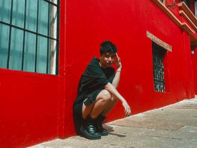 HoneiBun, agachada contra una pared de color rojo brillante, vestida con un vestido negro y botas, sosteniendo un teléfono en la oreja, con cabello corto y oscuro y una ligera sonrisa..