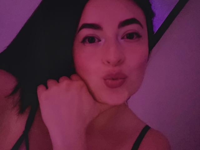 VaaleryStark with dark hair, pouty pink lips, and thick eyebrows, wearing a black top, holding her neck, in pink-purple lighting. Her tits are barely visible.