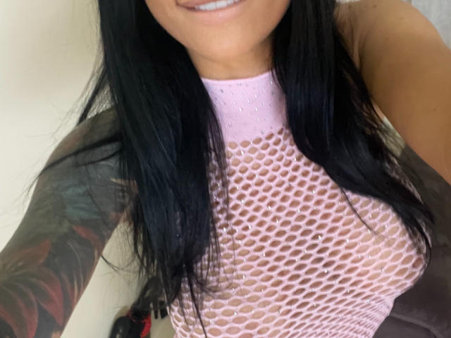 AndraD has black hair, smiling, wearing a pink mesh top that reveals her small tits and tattooed arm. Her pink top contrasts with her tan skin.