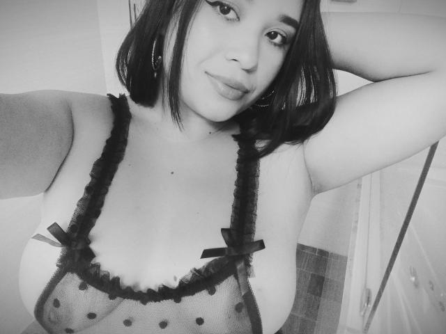 PamelaGoldwin with shoulder-length dark hair, wearing a sheer black lingerie top with polka dots and small bows, exposing her large, rounded tits, and a subtle smile.