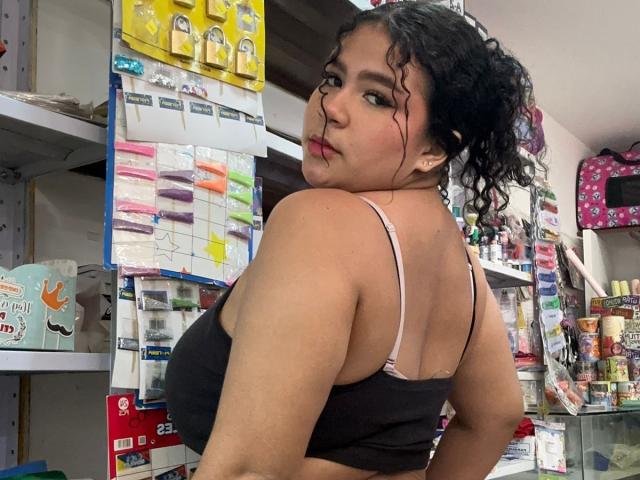 JulietaStark with curly black hair, brown skin, wearing a black sports bra, showing her big, round boobs, looking over her shoulder in a store.
