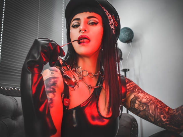 NinaBondQueen with dark makeup, tattoos, black leather gloves, hat, and red-tinted top, holding a whip near her red-lipsticked mouth, wearing a metal chain necklace.
