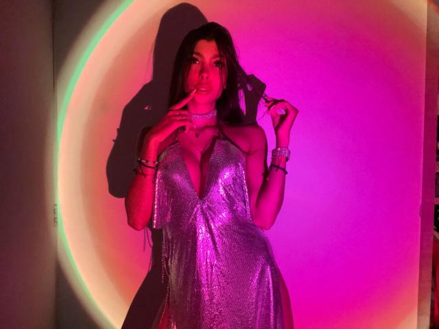 NatyJanssen in a sparkly purple dress with deep cleavage, holding a ring, pink and purple lighting, shadow on wall, long brown hair, seductive expression, finger to lips.