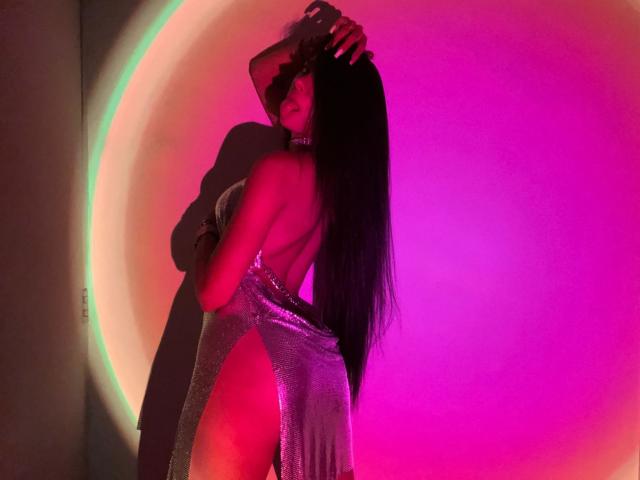 NatyJanssen in a silver, backless dress with a high slit, standing with a man, long black hair, pink and purple lighting, man's hand in her hair.