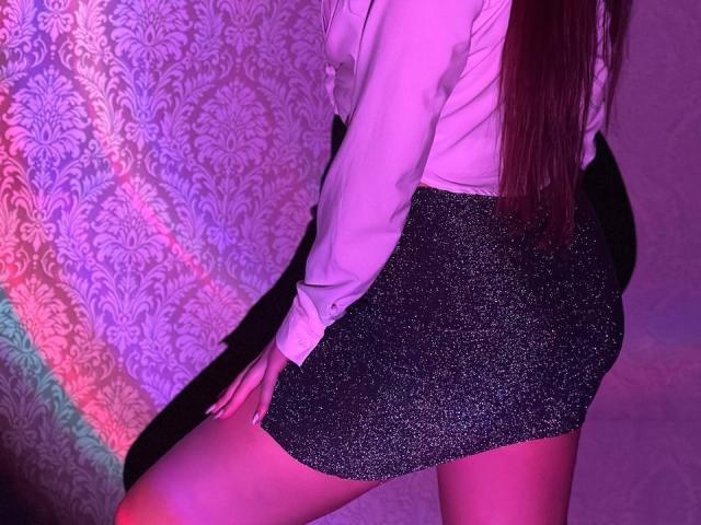 LisaAlisia's ass in a sparkly black mini-skirt, wearing a white long-sleeve shirt, with pink and purple lighting on her back and legs. Her hand rests on her thigh.