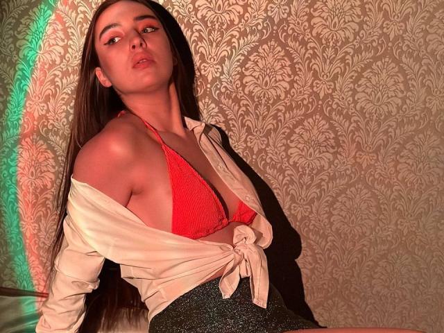 LisaAlisia with long brown hair, red lace bra, and tied white shirt, showing off her tits, sitting against a patterned wall with green and red light.