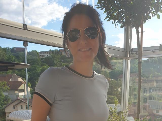 JoliDanielle with medium tits in a gray, short-sleeved, tight top, wearing aviator sunglasses, smiling, standing in a sunlit, glass-covered area, with a leafy tree to the right.