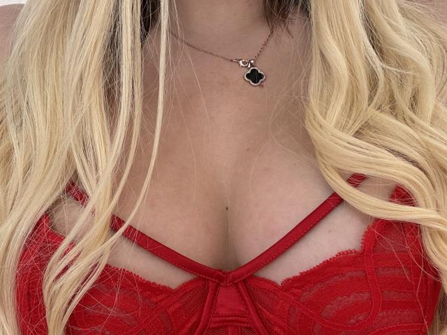 Close-up of NutaAlisson with blonde wavy hair, wearing a red lace bralette. She has fair skin, a small black pendant necklace, and visible cleavage. Textured lace and straps.