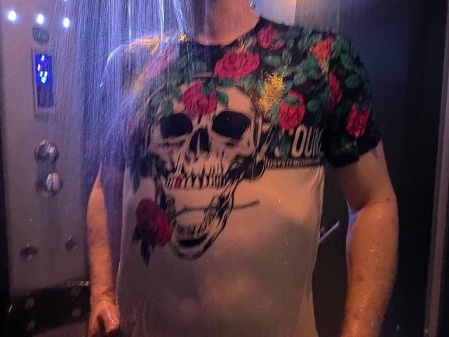 Svit with wet skin, wearing a skull and rose graphic tee, standing in a shower. Water drips down, showing a muscular, tattooed upper body. Svit's face is out of frame.
