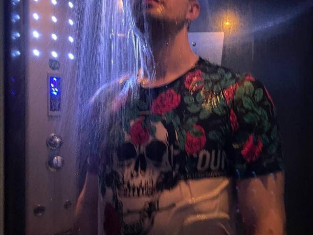 Svit with wet blonde hair, wearing a shiny skull and rose floral shirt, standing in a steamy shower with blue lights and water droplets on the glass.