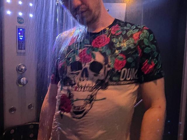 Svit stands under a shower, water dripping over his chest. He's wearing a wet, floral skull t-shirt, with colorful roses and text. His face is partially visible.