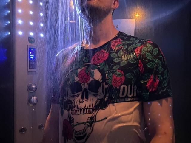 Svit with a skull and roses shirt, water spraying down, neon lights in background, wet shirt clinging to chest, arms slightly raised, facial features partially obscured.
