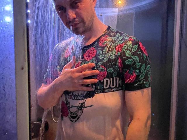 Svit, a muscular guy with a short beard, stands in a shower, water spraying on him, holding his chest with one hand, wearing a floral shirt, looking down.