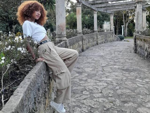 Cinthia Walker leunt tegen een stenen muur, gekleed in een wit shirt, beige cargobroek en witte sneakers, met krullend roodbruin haar, waarbij haar kleine borsten en slanke billen zichtbaar zijn..