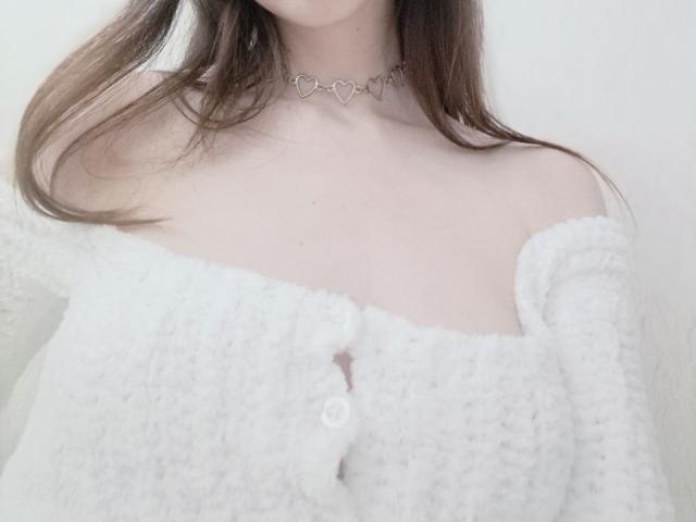 WollyMolly mit heller Haut, braunen Haaren, trägt einen weißen, strukturierten Pullover mit einem Träger, der von der Schulter abrutscht, und eine herzförmige Kette, die ihre Brüste zeigt. Ein weißer Knopf befindet sich in der Mitte des Pullovers..