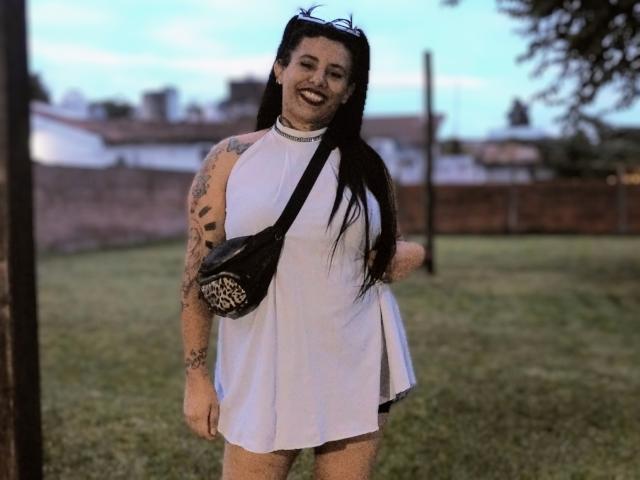 Yolisolange with black hair, tattoos on both arms, wearing a white sleeveless dress, black crossbody bag, and black cat ears, smiling in a grassy outdoor area.
