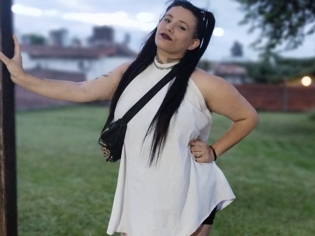 Yolisolange with black pigtails, white sleeveless dress, black cross necklace, black purse, and dark lipstick, standing with hand on pole, showing her ass slightly. Outdoor evening light.