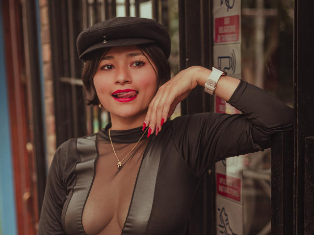 MiaLeblanc with short black hair, wearing a black cap, sheer black top with deep cleavage, red nails, gold necklace, and wristwatch, sticking out pink tongue, sexy.