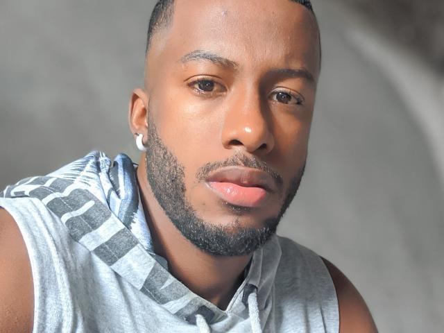 NegroEbano with dark brown skin, short black hair, and a trimmed beard, wearing a grey sleeveless shirt, has shiny pink lips and a small white earring in his left ear.