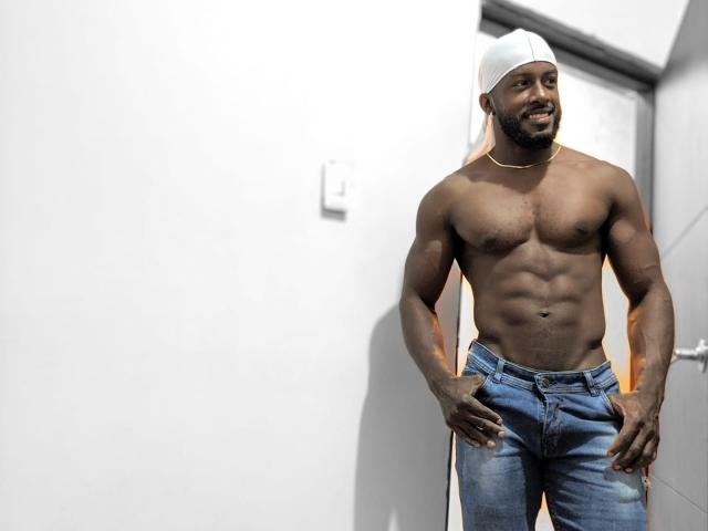 Muscular NegroEbano with dark skin, wearing a white backwards cap and blue jeans, stands shirtless, showing defined abs and a gold chain, smiling confidently. Hands in pockets.
