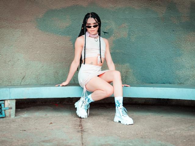 AbbyR with long braids, sunglasses, white crop top, high-waisted skirt, and white boots, sitting on blue bench, showing her ass, with a pink choker, and white socks.