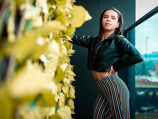 AbbyR with dark hair, wearing a green leather crop top and striped high-waist pants, leaning against a yellow leaf wall, hand on hip, looking confident.