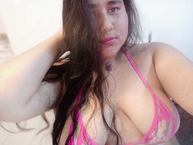 ZoeGiil with long brown hair, pink lace bra, showing large boobs, fair skin, pink lipstick, purple necklace, hand in hair, slightly blurred, soft lighting, looking at camera.