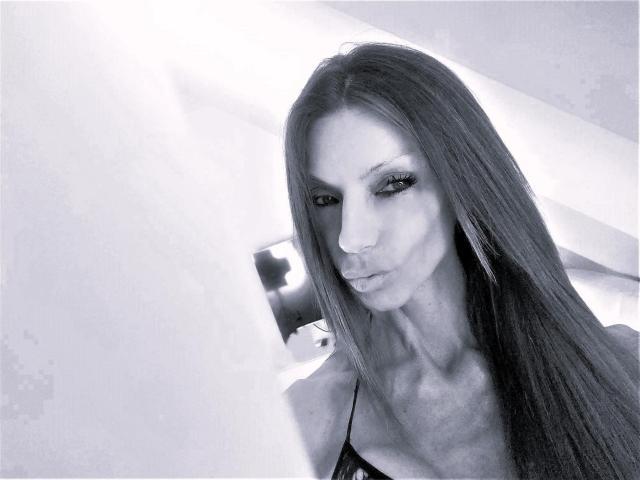 MonikaPrize with long straight dark hair, pouty lips, and dark eyeliner, wearing a black lace top, showing her tits, in a high-contrast black-and-white photo.