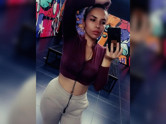 VioletaStone taking a mirror selfie, wearing a maroon zip-up crop top and white sweatpants, with her arm raised, showing her flat stomach and small, perky tits.
